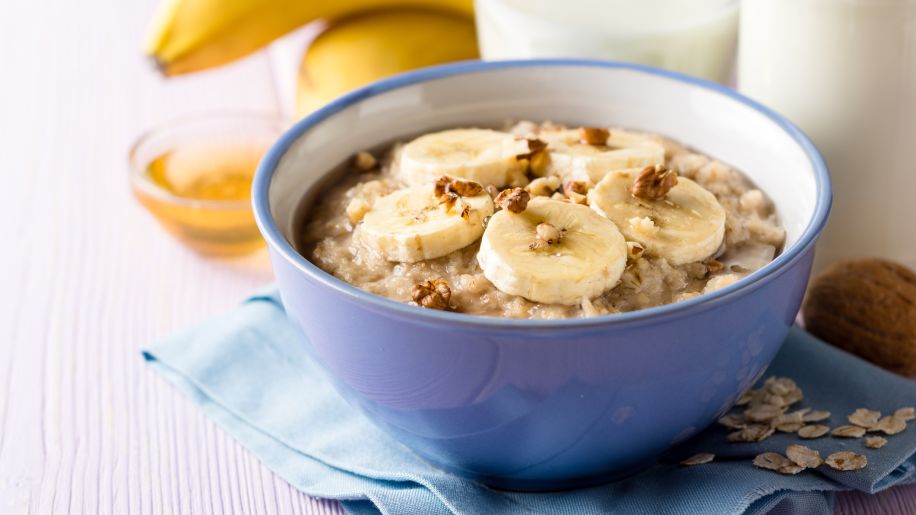 A bowl of oatmeal topped with sliced bananas and walnuts, with honey, bananas and glasses of milk in the background