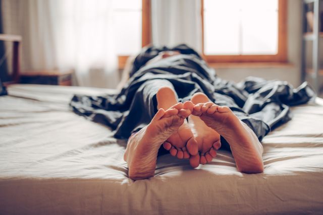 A smiling couple snuggles under the sheets. They know healthy, monogamous sex is good for anxiety, stress and your health.