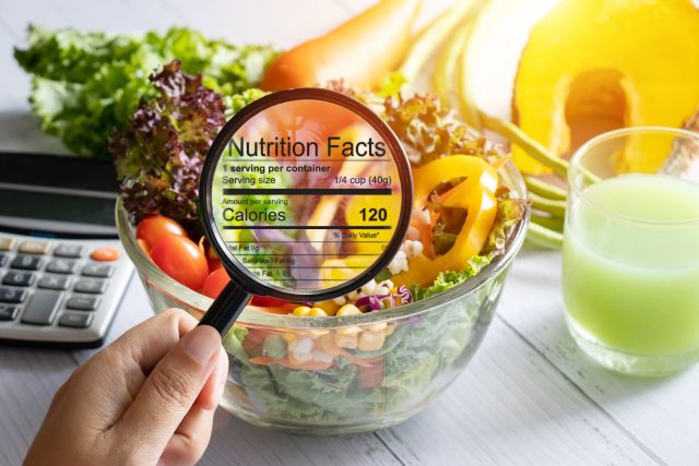 A consumer holds a magnifying glass up to a healthy salad, with a nutrition facts label superimposed over the salad