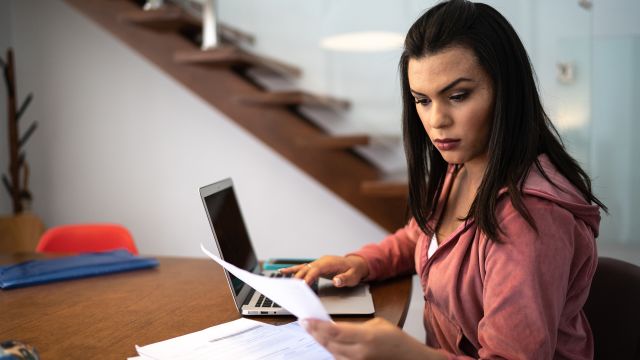 Transgender woman reading papers while working on a laptop.