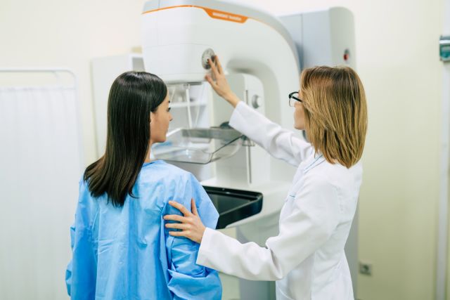 A patient in a blue gown faces a mammogram machine, while a healthcare provider stands by.