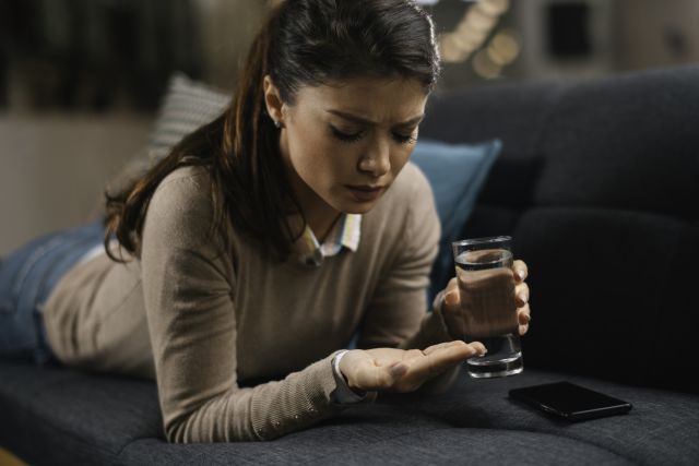a women woman lies on her couch about to take a pill with a smartphone nearby