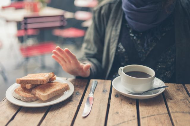 a woman sitting at an outdoor cafe drinking coffee declines to eat bread because she has celiac disease
