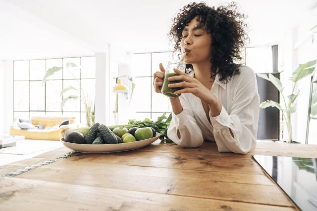 African American woman enjoying a green smoothie with a bowl of green fruits and vegetables in front of her