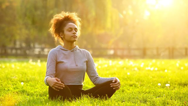A woman mediates outside in the sunshine. Stress relief and mindfulness can be helpful to people with psoriasis.