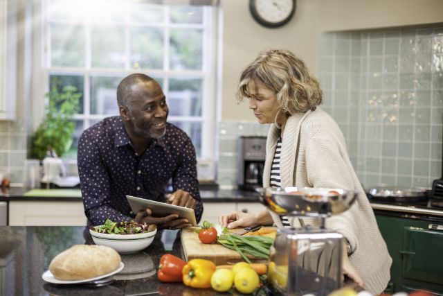 A middle aged man and woman look at a computer tablet and discuss a recipe, while surrounded by fresh fruits and vegetables in a sunny kitchen.