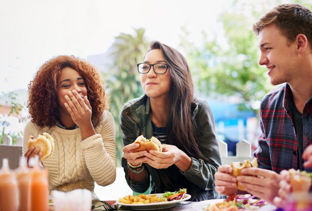 A group of friends consisting of a Black woman, an Asian American woman, and a white man, enjoy a meal of hamburgers together at a restaurant