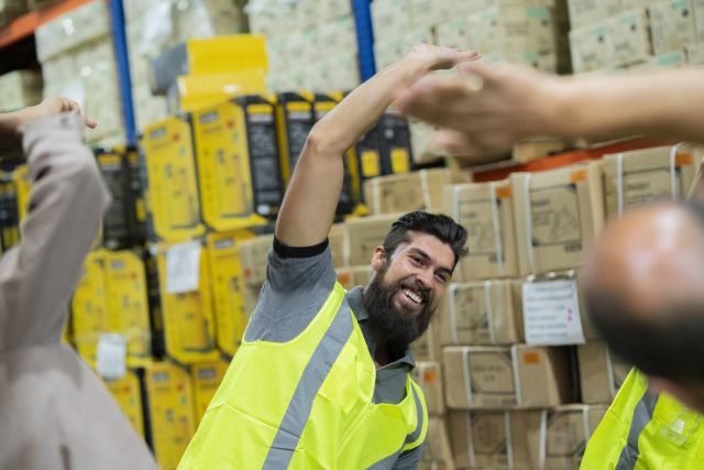 a bearded young man wearing a yellow vest performs stretches as part of a workplace exercise routine in a warehouse work setting