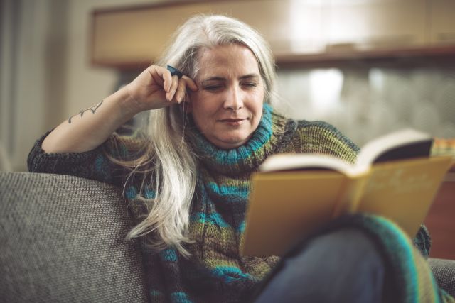A middle aged white woman with gray hair and a tattoo on her arm sits in a couch reading a book