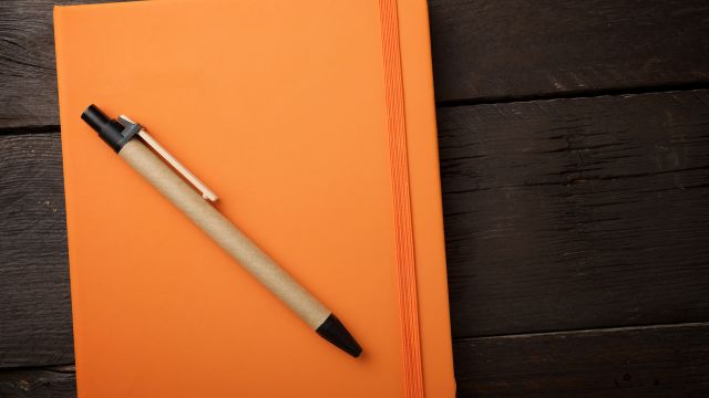 A pen lays on top of an orange notebook.