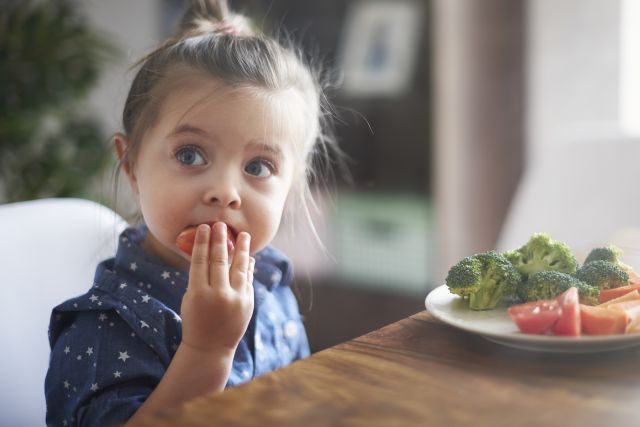 a young White child sits at a table in front of a plate of chopped vegetables, placing a tomato piece in her mouth