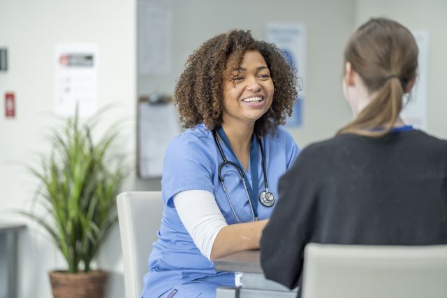 a young Latina nurse smiles and consults with her patient, a female who is in the foreground