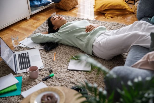 view of woman lying on the floor on her back with her eyes closed, surrounded by her laptop, notebooks and coffee