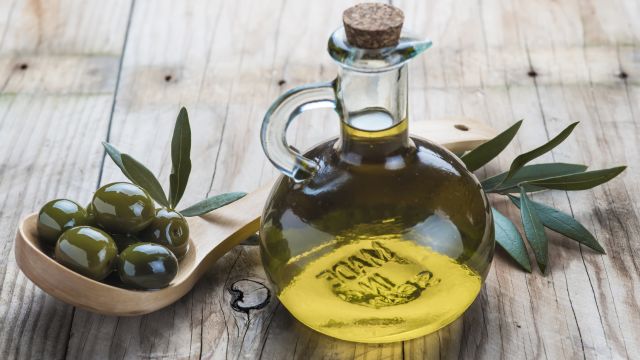 A glass bottle of olive oil and a wooden spoon with  olives on a table