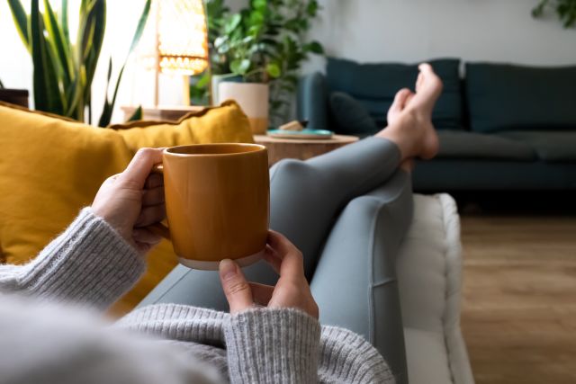 a woman reclines on a couch, holding a mug of tea; we see the tea from the perspective of the woman, with the mug of tea in the foreground and her feet in the background