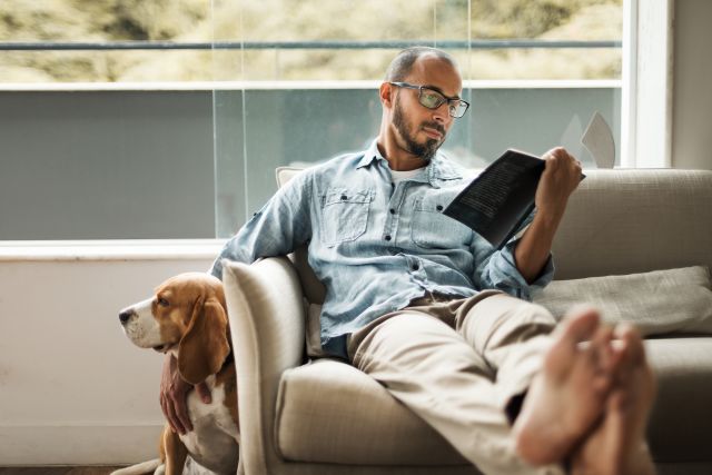 Bearded man comfortably sitting on a coach to de-stress by reading a book and holding his dog