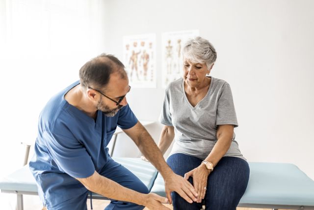 Senior woman with osteoarthritis sitting on an exam table while a male physical therapist examines her knee
