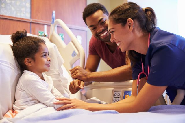 Smiling pediatrician leaning on hospital bed of happy young girl, her father also leaning in and smiling