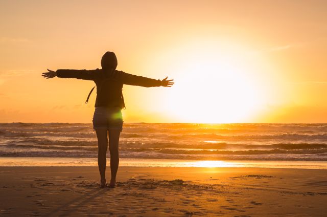 rear view of a woman standing on a beach with arms outstretched overlooking the sunset  over the water