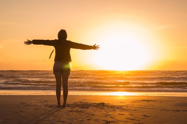 back view of someone standing on a beach with arms outstretched overlooking the sunset over the water