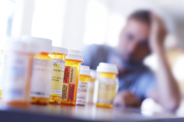 closeup of an assortment of prescription pill bottles with a distressed man staring at them in the blurry background