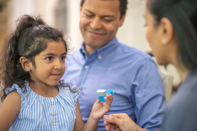 A young Latina girl meets with a healthcare provider to learn how to use her inhaler for her childhood asthma symptoms with her caregiver nearby