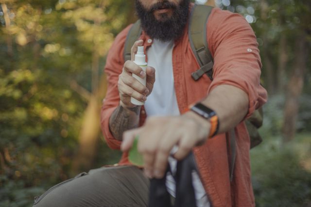 man with a beard in a wooded area spraying mosquito repellent onto his arm to prevent zika virus