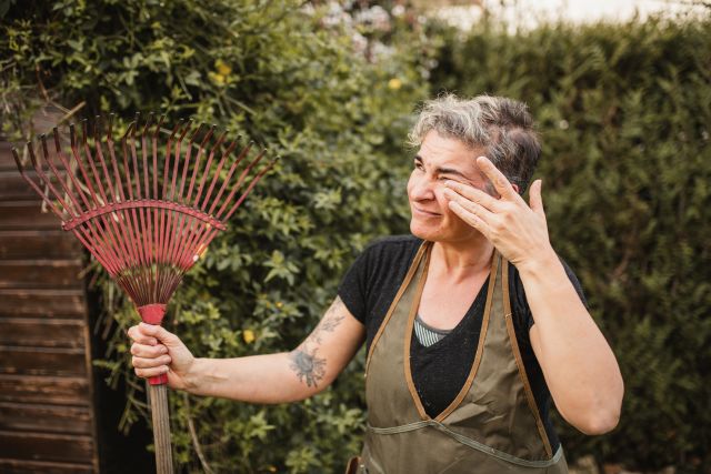 a middle aged woman with a tattoo on her arm rubs her eyes as she works in her garden because she has seasonal allergies