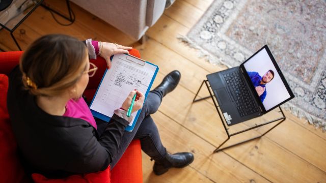 A therapist in a home office speaks with a patient during a telehealth therapy appointment.