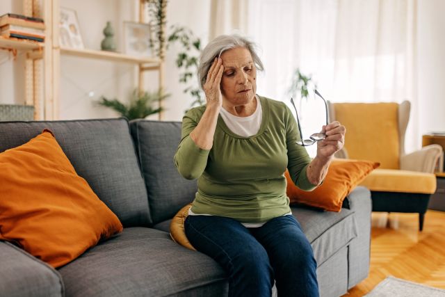 An older white woman sits on her coach holding her head and removing her glasses. Her vision is blurry and she has a headache. Could she be having a stroke?