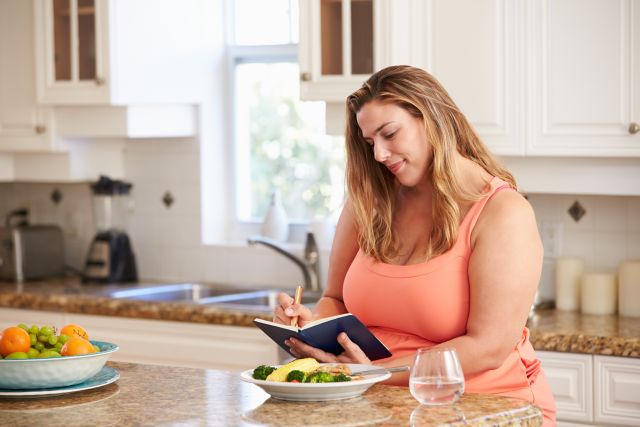 an overweight white woman in a pink tank t-shirt sits at her kitchen counter and smiles as she makes an entry into her food diary to help track which foods cause heartburn