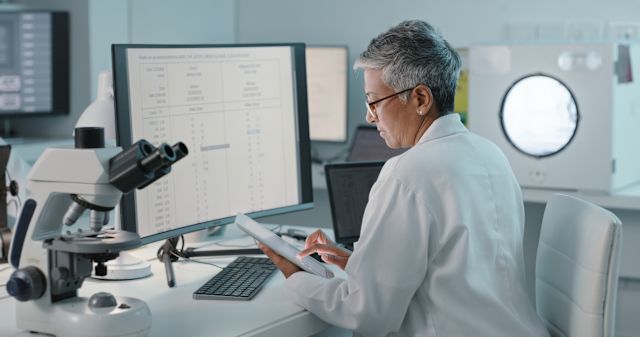 a middle aged female South Asian scientist with short gray hair sits in a science laboratory reviewing data on COVID vaccine development on a computer and tablet screen  