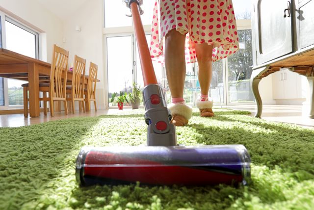 A closeup shot of the front end of a vacuum while a woman wearing house slippers and a red polka dot dress vacuums dust off of a green shag rug