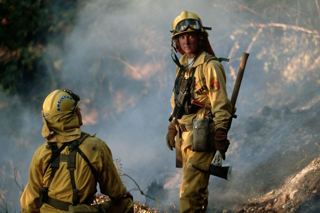 Two firefighters, a man and a woman, in full yellow firefighting turnout gear, stand amid the smoky remnants of a large forest fire