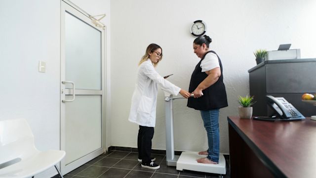 A woman stands on a scale during an appointment with a healthcare provider. Weight and height measurements are used to calculate body mass index (BMI) and diagnose obesity.