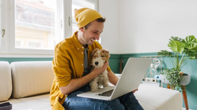 A young man holds his dog on his lap while he participates in an online support group for people with HAE using his laptop computer.
