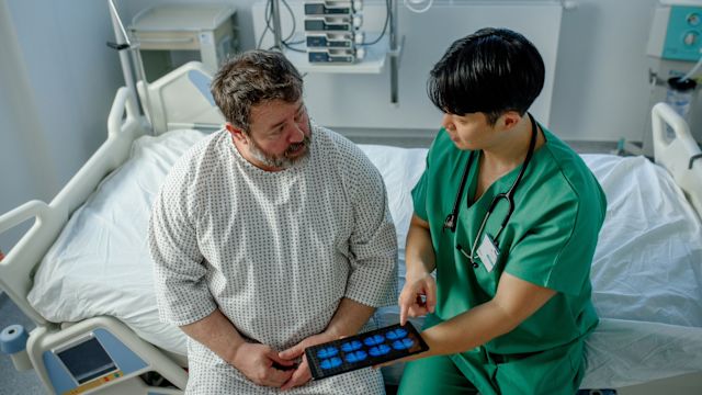A neurologist reviews the results of a brain imaging scan with a male patient in a hospital room.