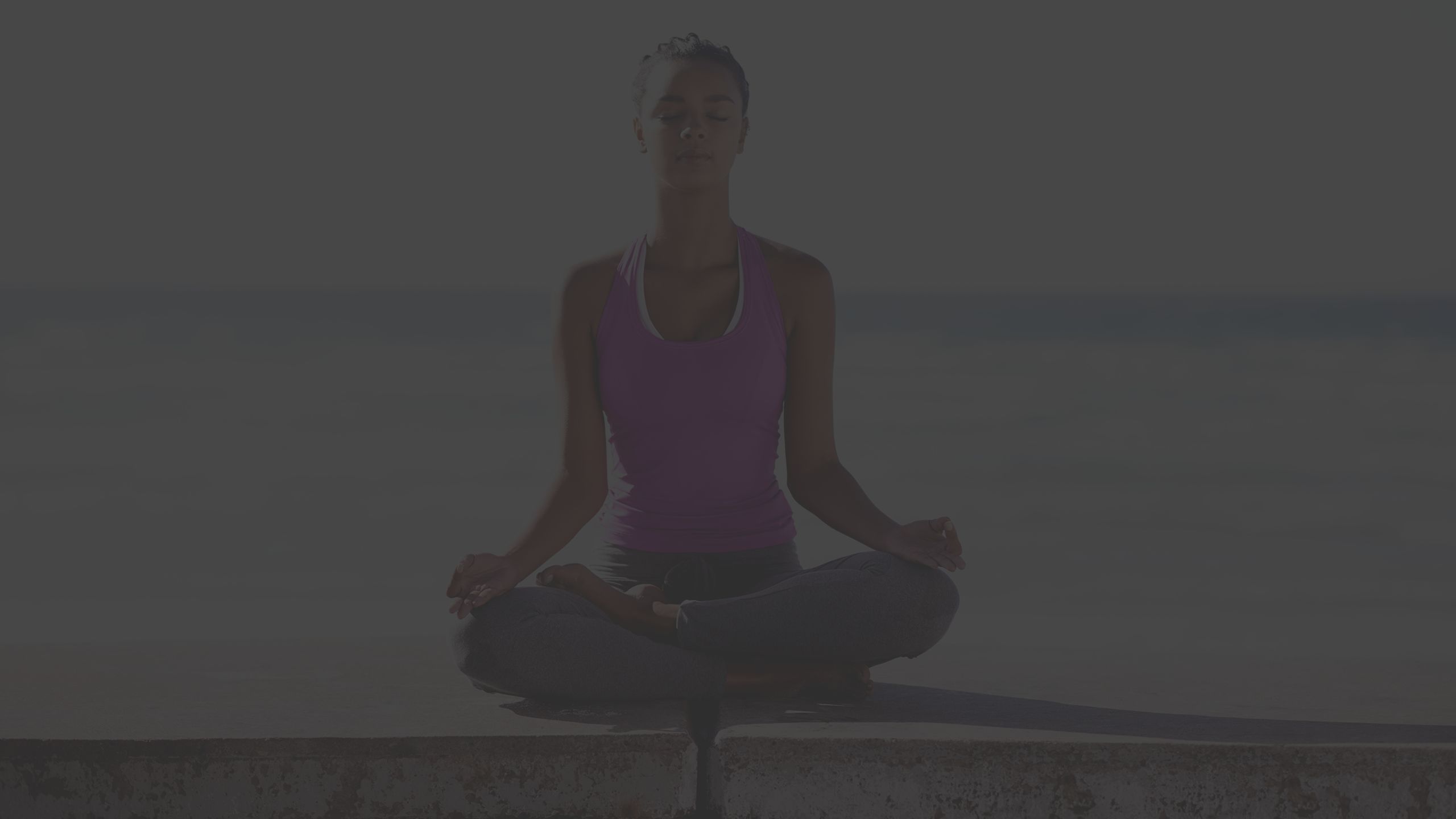 young woman sitting on a wall practicing yoga with the beach behind her