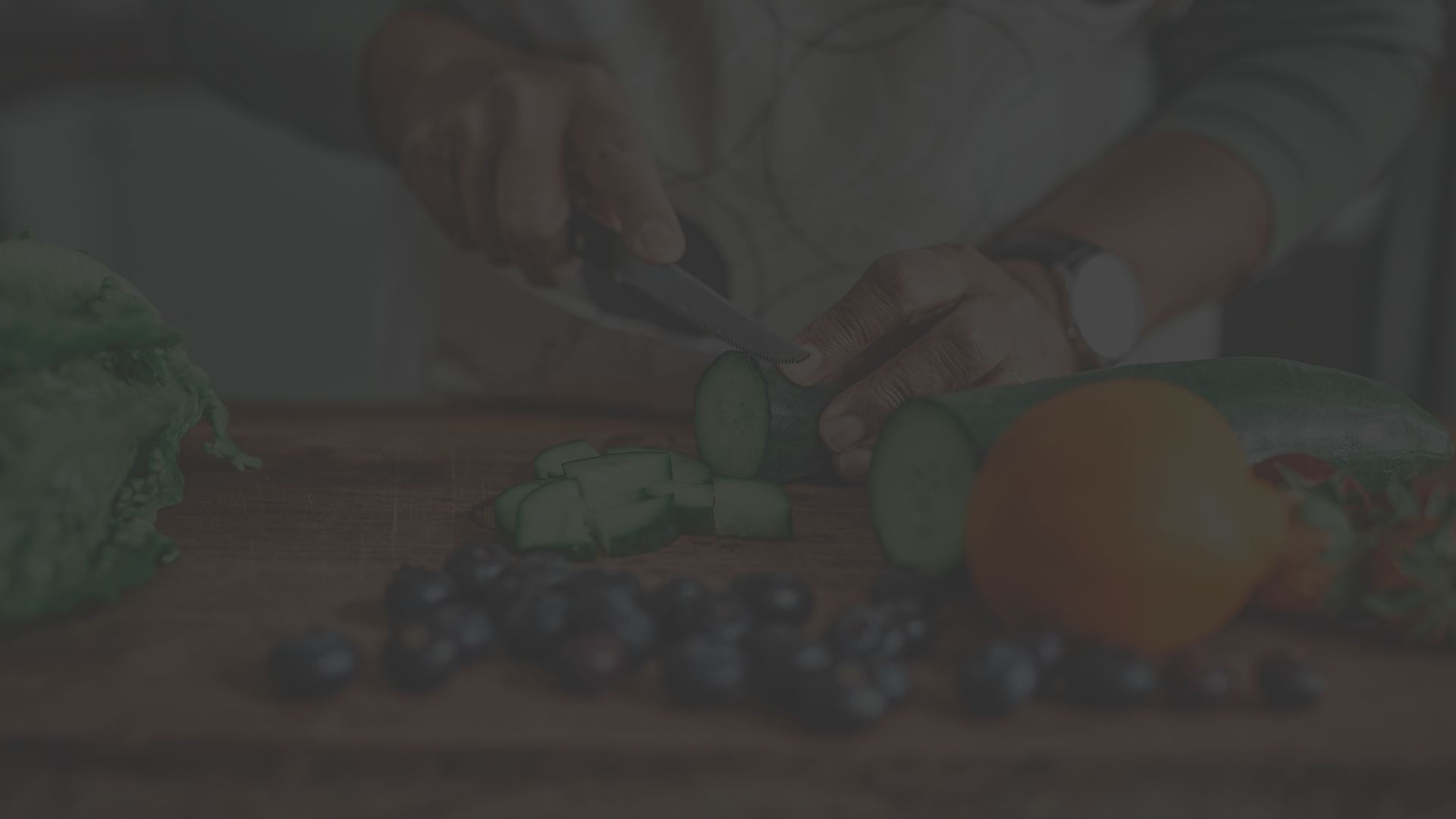 View of an unidentifiable man cutting up cucumber on a cutting board with other vegetables and fruits on the counter