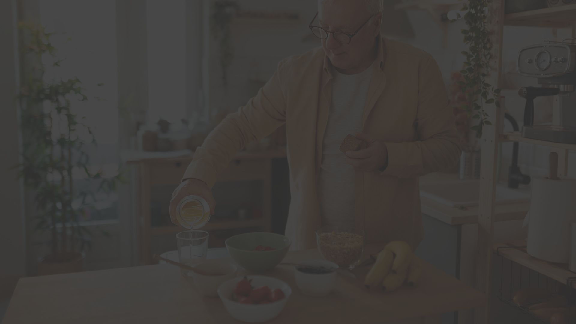 an older man with white hair and glasses pours fresh orange juice into a glass in his kitchen with a bowl of strawberries, blueberries and bananas on the counter