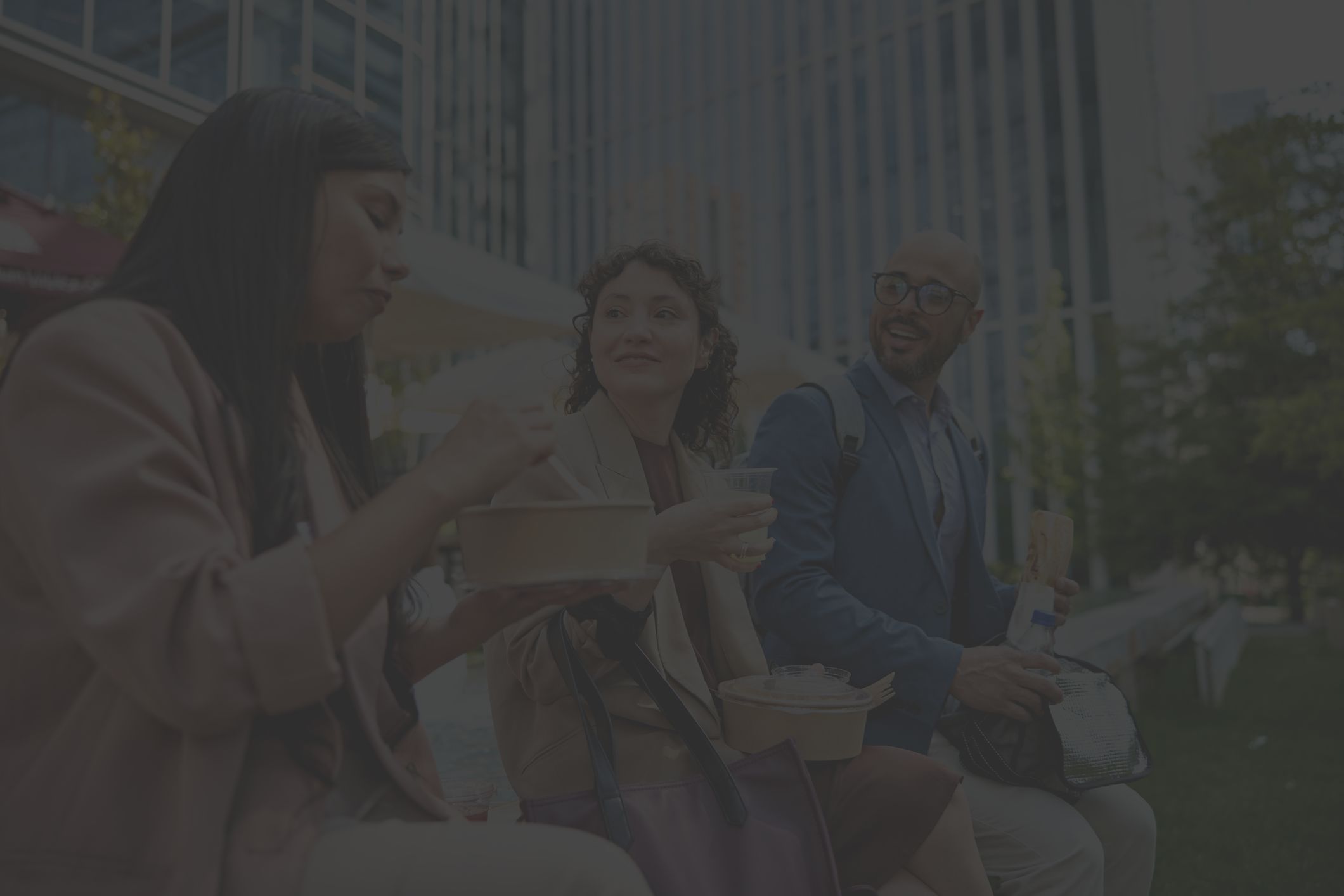 three office workers, a Latina woman, a white woman, and a Black man, enjoy eating lunch outside on a spring day
