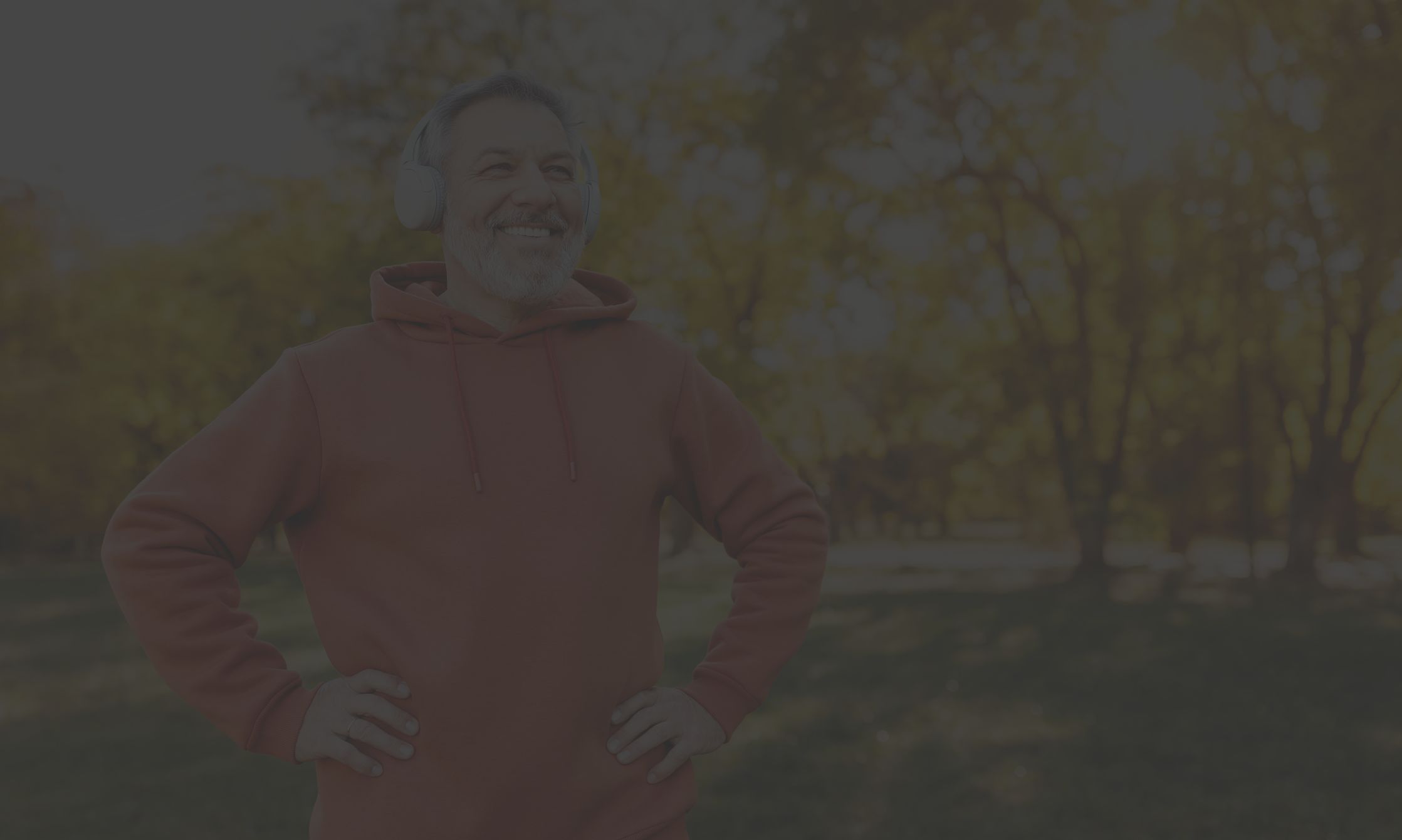 a smiling middle aged Hispanic man with a beard wears earphones and a red sweatshirt as he gets ready to exercise outdoors in a park