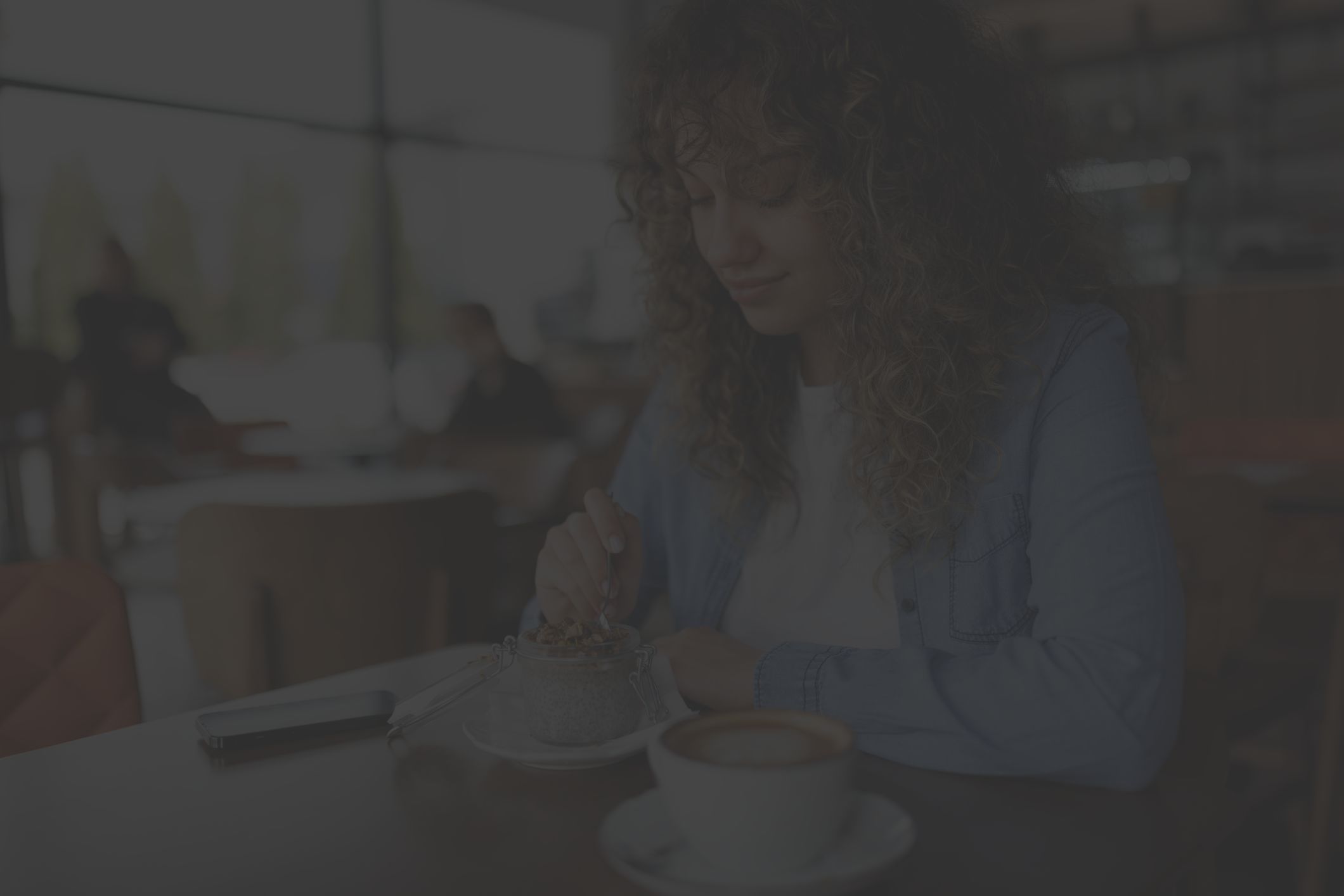 young, curly-haired woman eating chia seed pudding and drinking coffee at a cafe