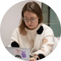 A young woman with glasses and a patterned sweater works on a laptop.