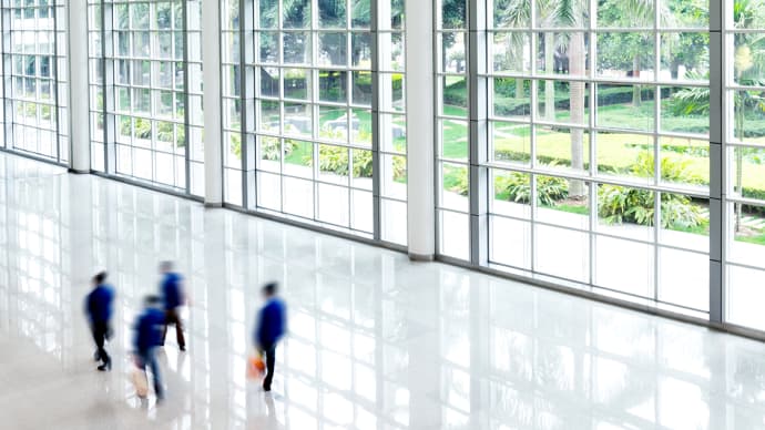 Motion-blurred figures walk across a bright, polished hall with floor-to-ceiling windows overlooking a green garden.